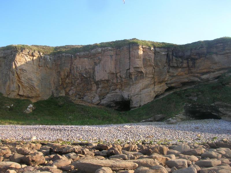 A photograph of a stony shoreline leading to a grassy slope and a cave set in a dramatic landscape of vertical cliffs